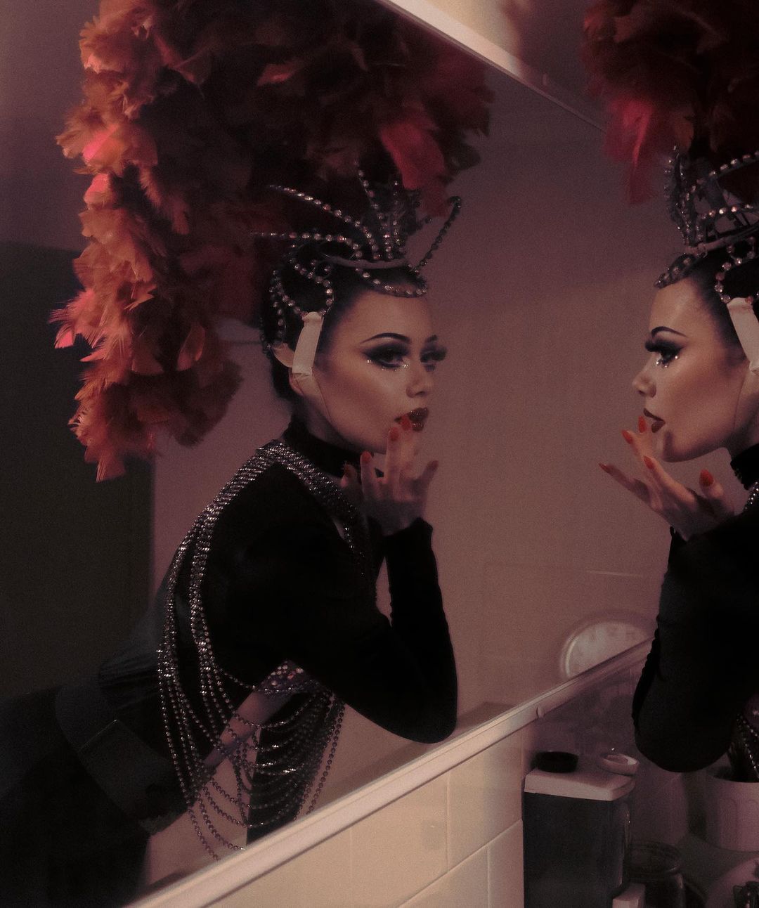 A woman in elaborate costume poses in front of a mirror, showing off her makeup, feathers, and beaded details in an indoor setting.