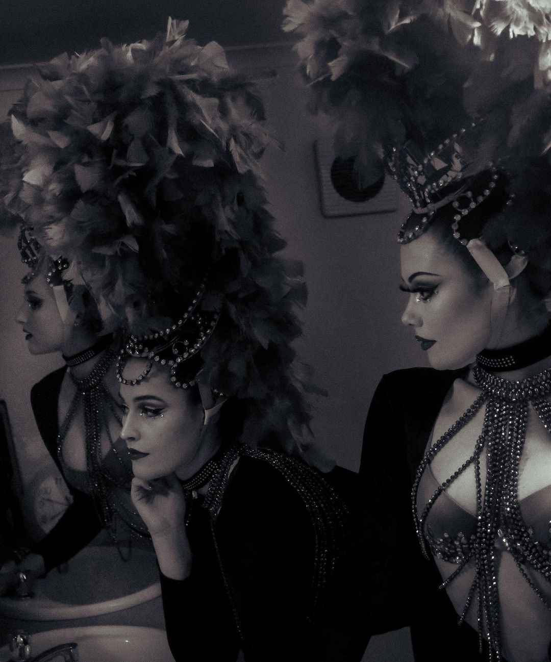 Portrait of an adult female wearing a feather headpiece and beaded top, looking at herself in a mirror in an indoor setting.