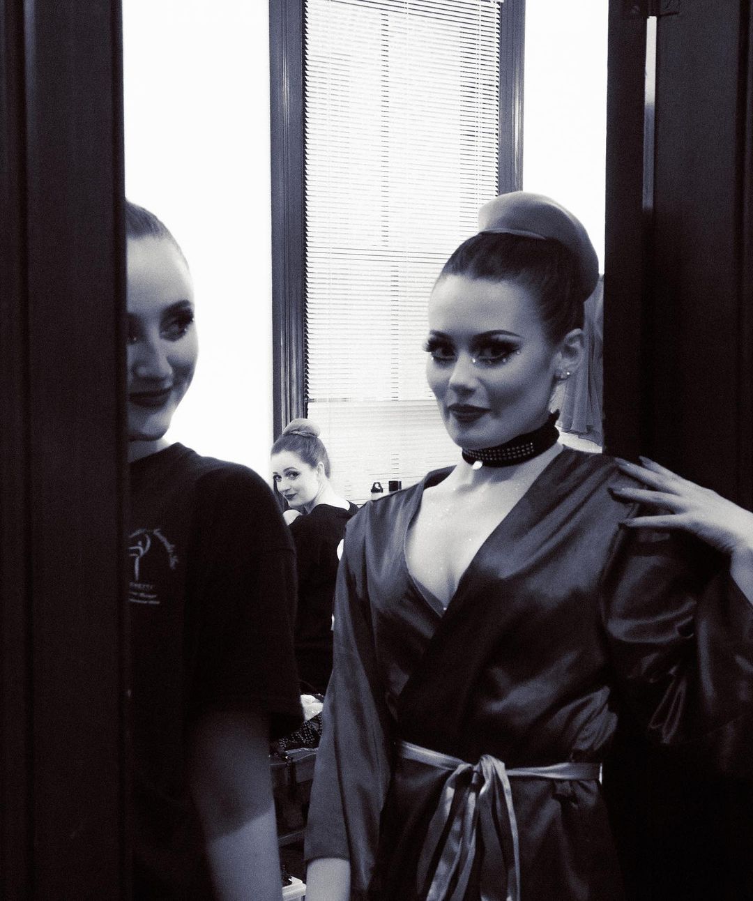 Two women pose indoors; one is in a robe and makeup, the other looks on near an open door with a third woman visible in the background.