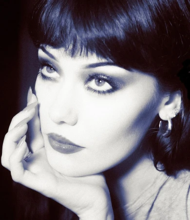 Close-up portrait of a woman with dark hair and bangs resting her chin on her hand, featuring bold eye makeup.