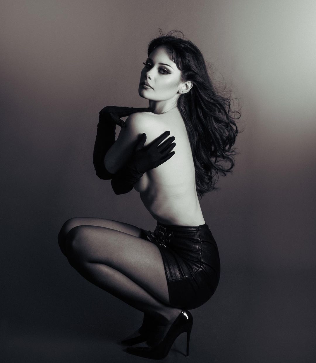Black and white portrait of a woman in a short leather skirt, gloves, and high heels, posing in a studio with soft lighting and arms crossed.