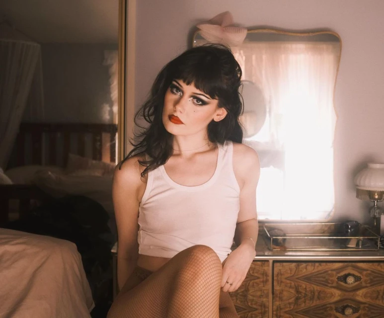 Portrait of a young woman with dramatic makeup and a white top sitting in a bedroom with a bed and vanity in the background.