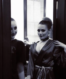 Monochrome backstage portrait of a woman wearing a satin robe and choker, framed by doorway edges with another woman visible in reflection