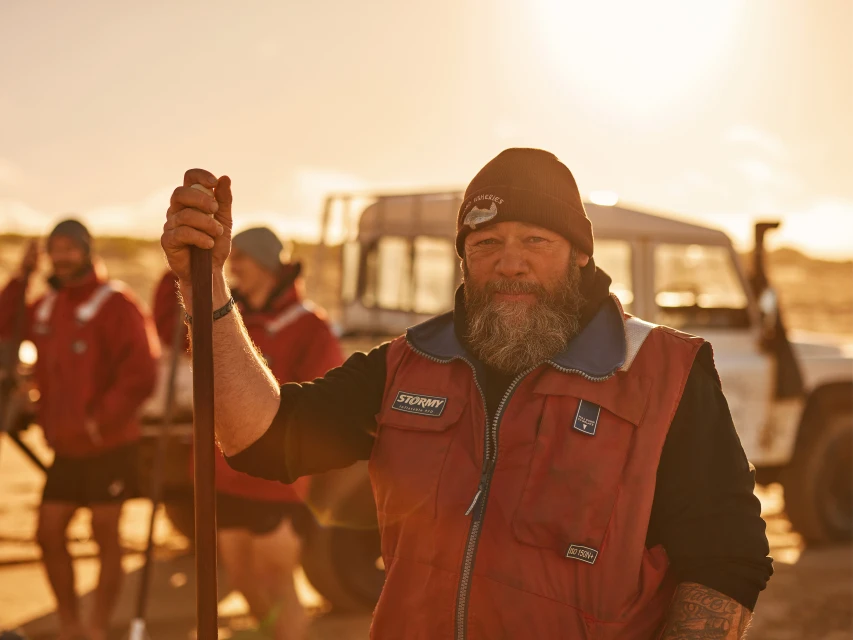 Portrait of bearded man in beanie and red vest on a beach with other people and a vehicle in the background during daytime.