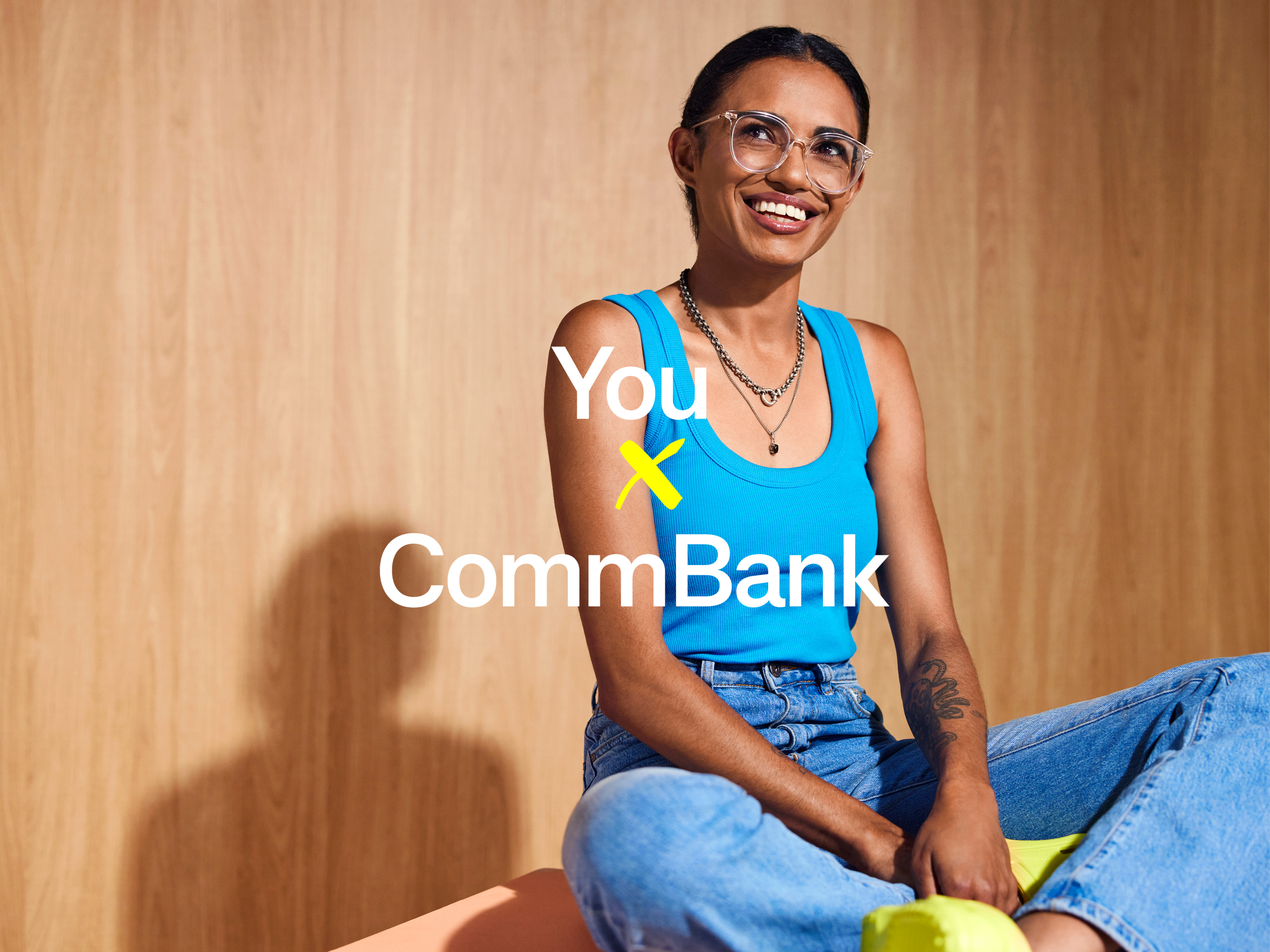 A smiling young woman with glasses and a blue tank top sits in front of a wood-paneled wall. The CommBank logo is overlaid.
