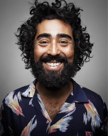 Portrait of smiling man with dark curly hair and a beard wearing a patterned navy blue shirt against a grey background