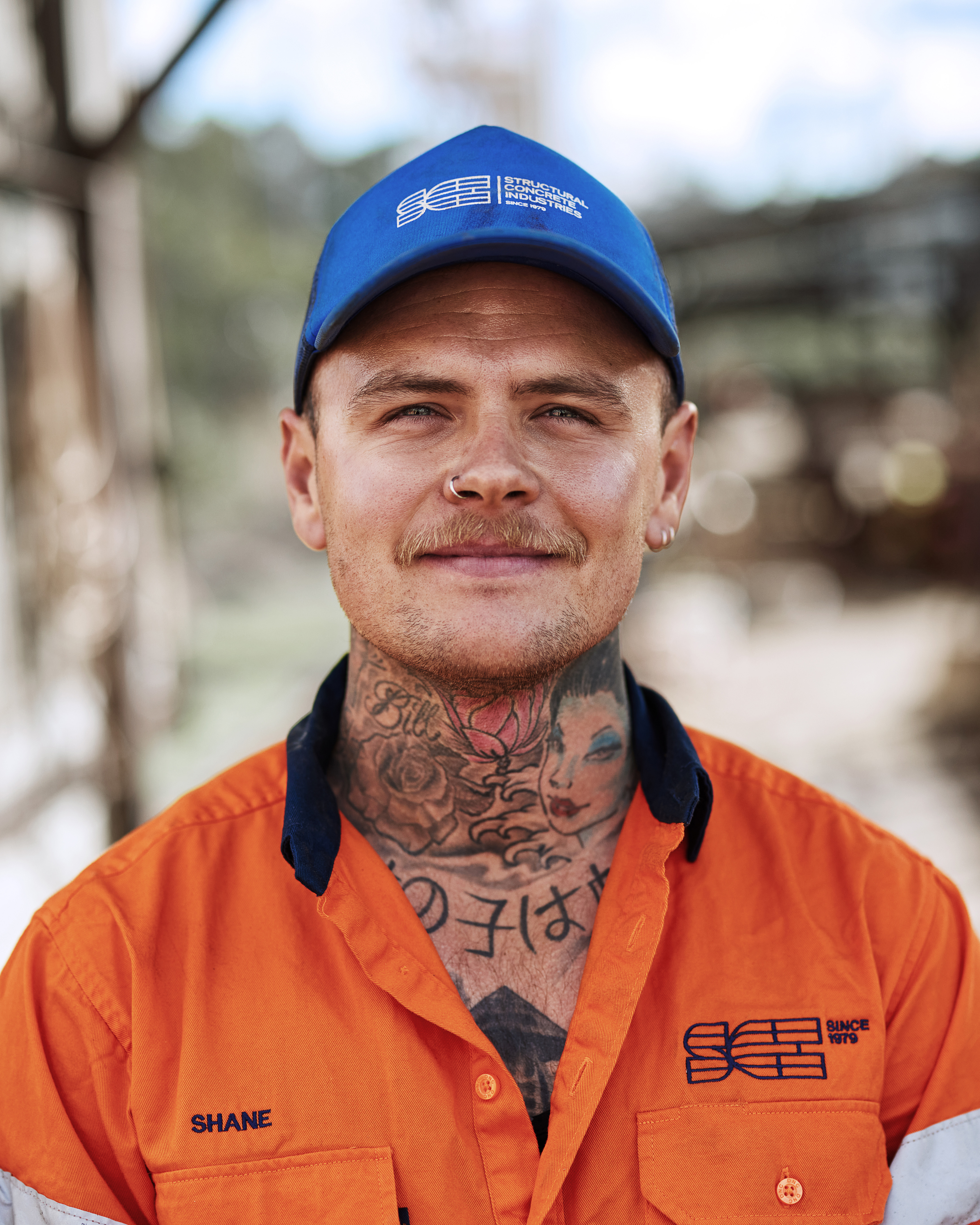 Close-up portrait of a tattooed man in an orange work shirt and blue cap, smiling slightly, set against an outdoor background.