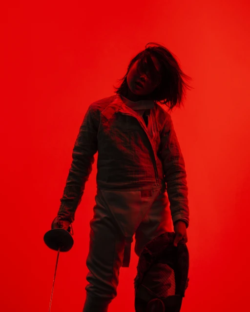 Fencer in red light stands with sword, wearing protective gear, creating a dramatic silhouette effect against the vibrant backdrop.