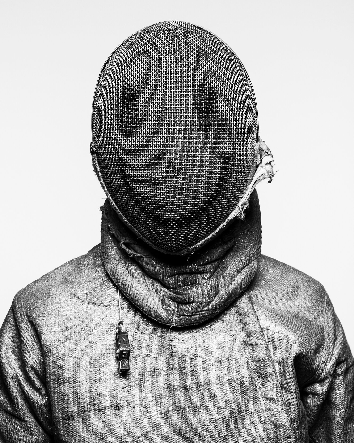 A person in a fencing uniform with a smiley face on the fencing mask stands against a white background in a black and white studio shot.