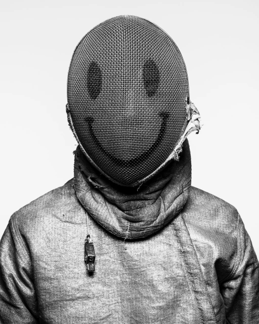 A person in a fencing uniform with a smiley face on the fencing mask stands against a white background in a black and white studio shot.