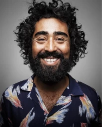 Portrait of smiling man with dark curly hair and a beard wearing a patterned navy blue shirt against a grey background