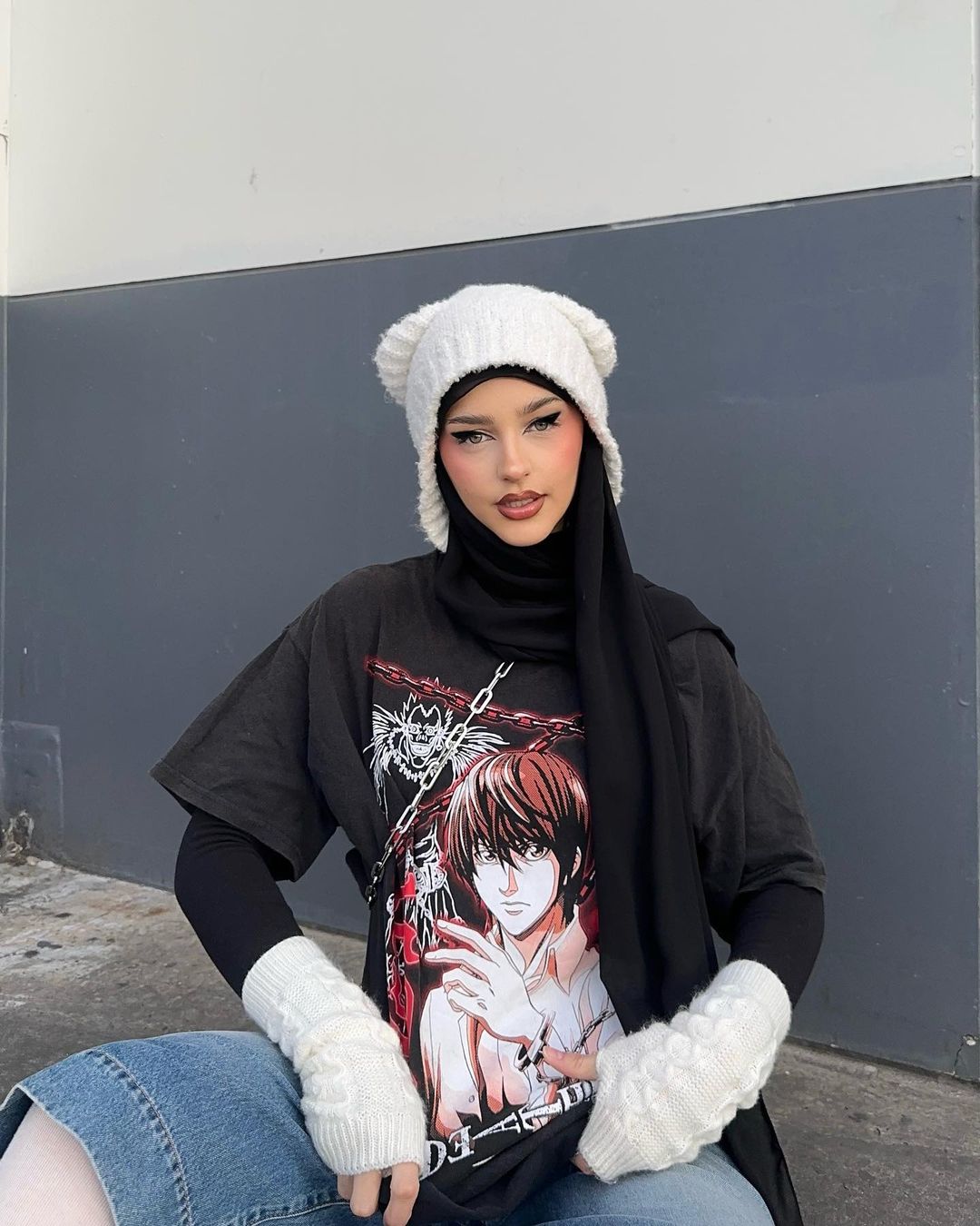 Adult female in an anime t-shirt, knit hat with bear ears, and arm warmers poses against a grey wall, wearing a black hijab.