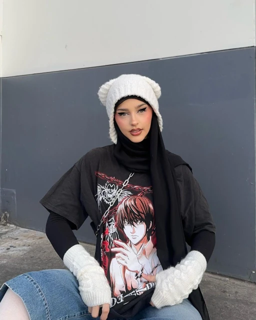 Adult female in an anime t-shirt, knit hat with bear ears, and arm warmers poses against a grey wall, wearing a black hijab.