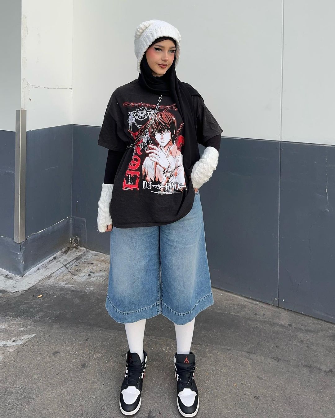 Young woman in trendy streetwear posing against a grey wall, wearing a t-shirt, denim culottes, sneakers, and a knit beanie.
