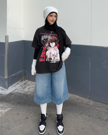 Young woman in trendy streetwear posing against a grey wall, wearing a t-shirt, denim culottes, sneakers, and a knit beanie.