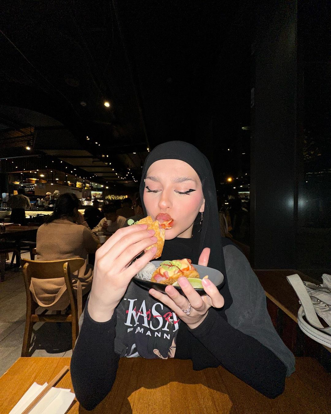 Adult female in a headscarf dines at a restaurant, holding a plate with food and a taco, with restaurant patrons in the background.