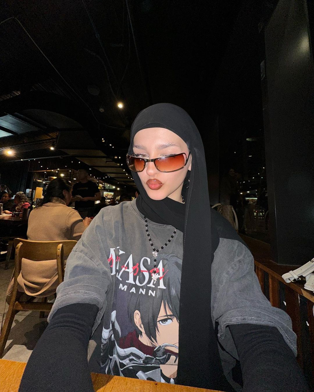 Portrait of a woman wearing a black hijab and sunglasses, posing indoors. The background features warm lighting and a restaurant setting.