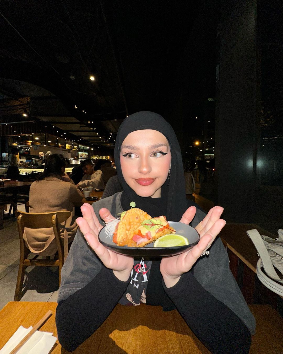 Woman in hijab smiles while holding a plate with two tacos in a dimly lit restaurant, with other patrons visible in the background.
