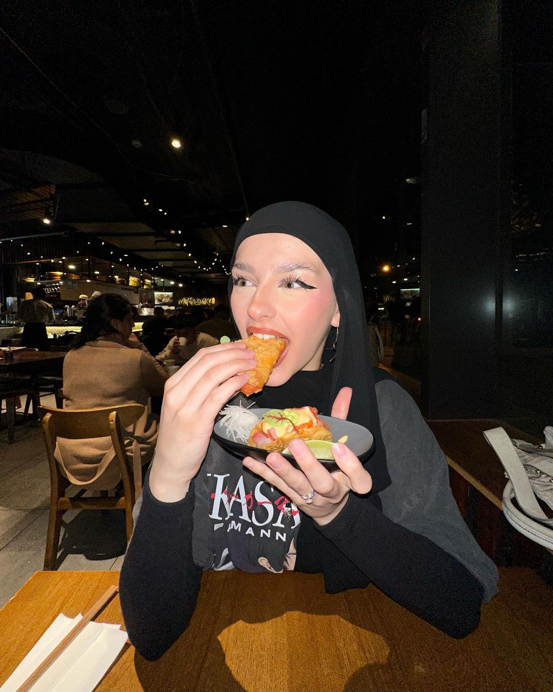 A woman in a black hijab and top is eating a taco off a plate at a restaurant table. There are other people and interior lighting visible.
