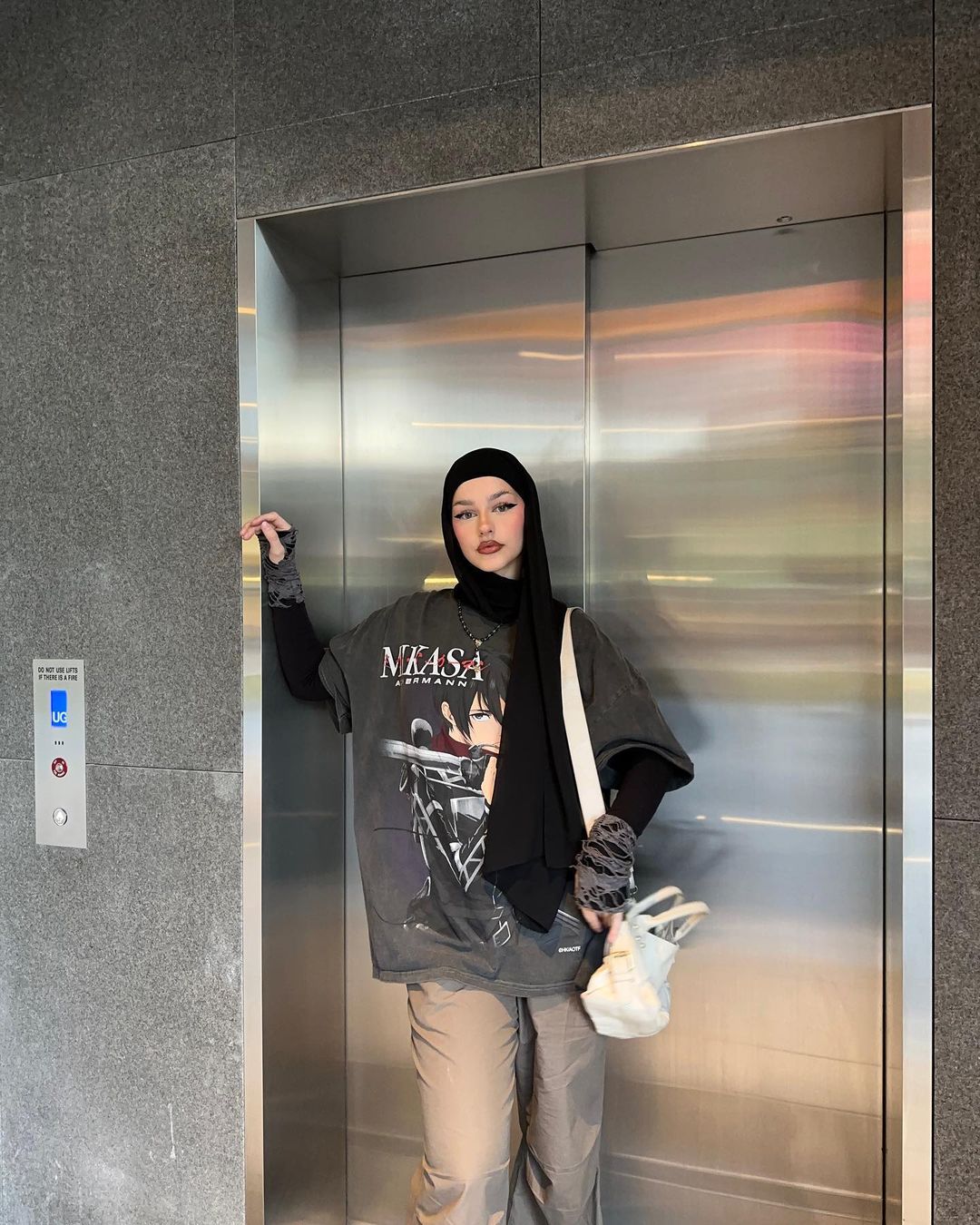 A stylish woman in a black hijab and oversized graphic tee poses inside an elevator with cool gray and silver interior panels.