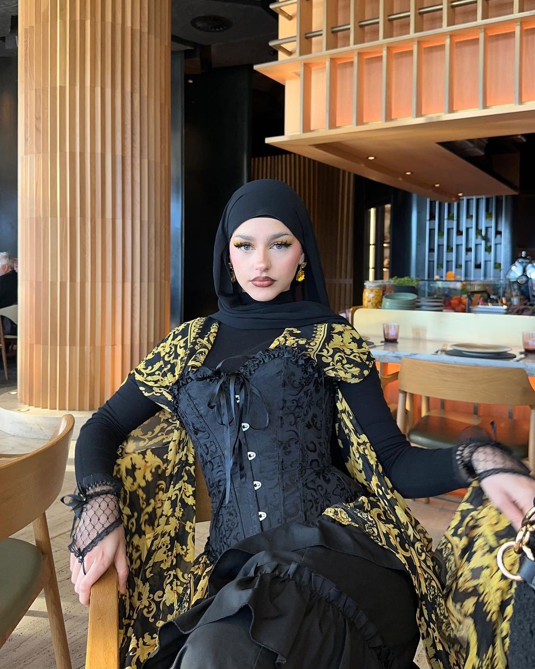 Portrait of a fashionable adult woman indoors wearing a black headscarf, black top, and a black and gold patterned corset.