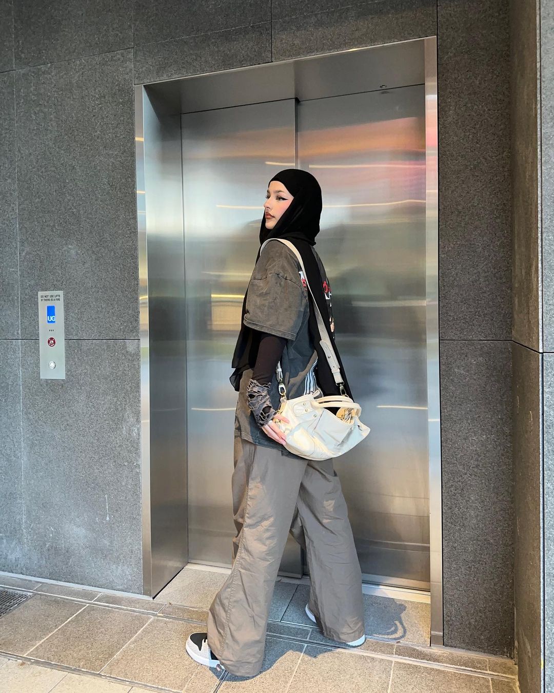 A woman in wide leg pants, a dark shirt, and a black hijab stands in front of a metal elevator looking to the side. The photo is indoors.