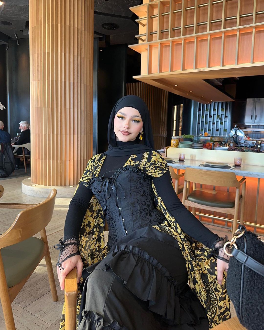 Adult female in hijab and black dress sitting in a chair inside a restaurant with natural lighting, fair skin and green eyes.