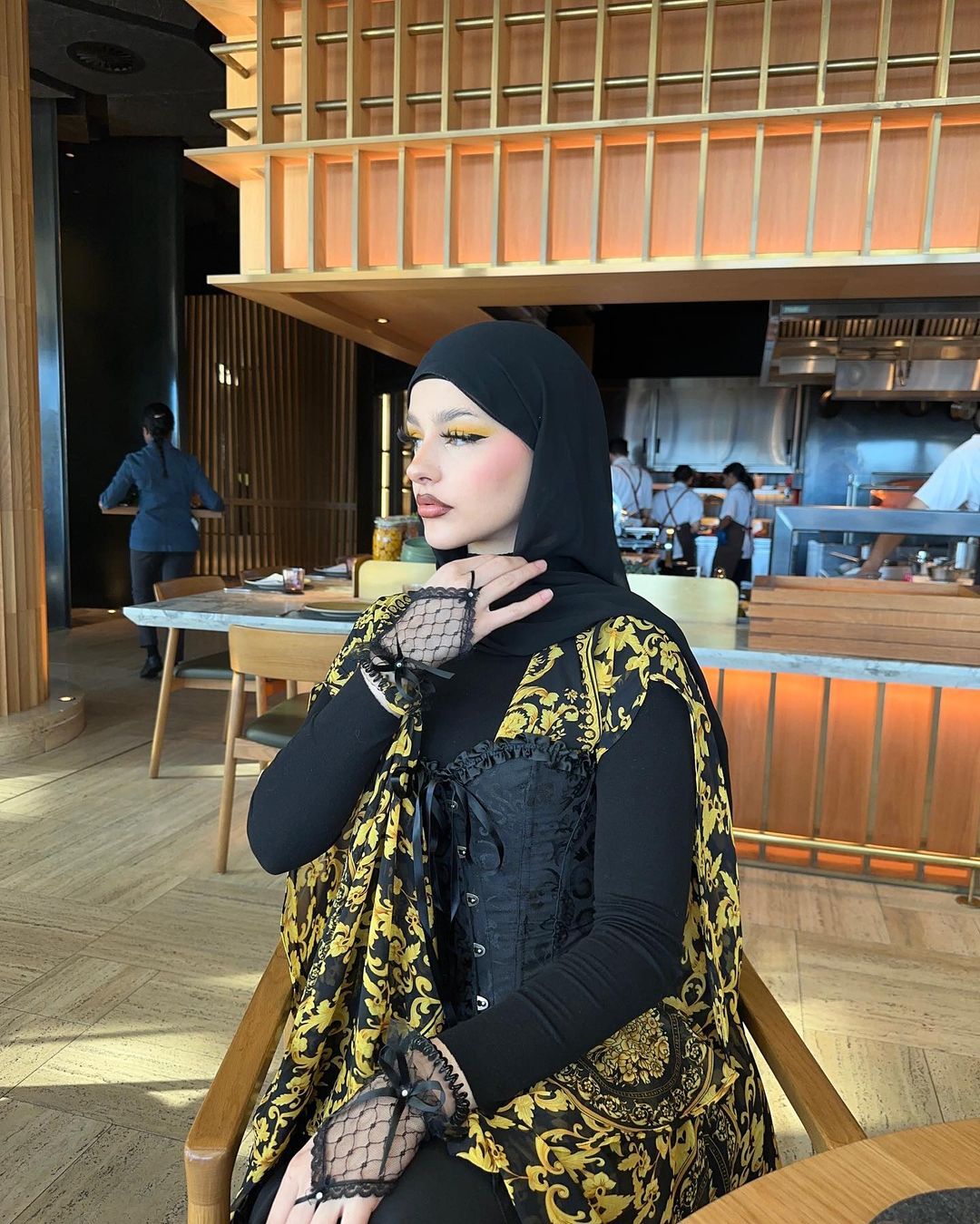 Woman in black hijab and patterned shawl sitting in restaurant, looking away. Modern interior design and natural light.