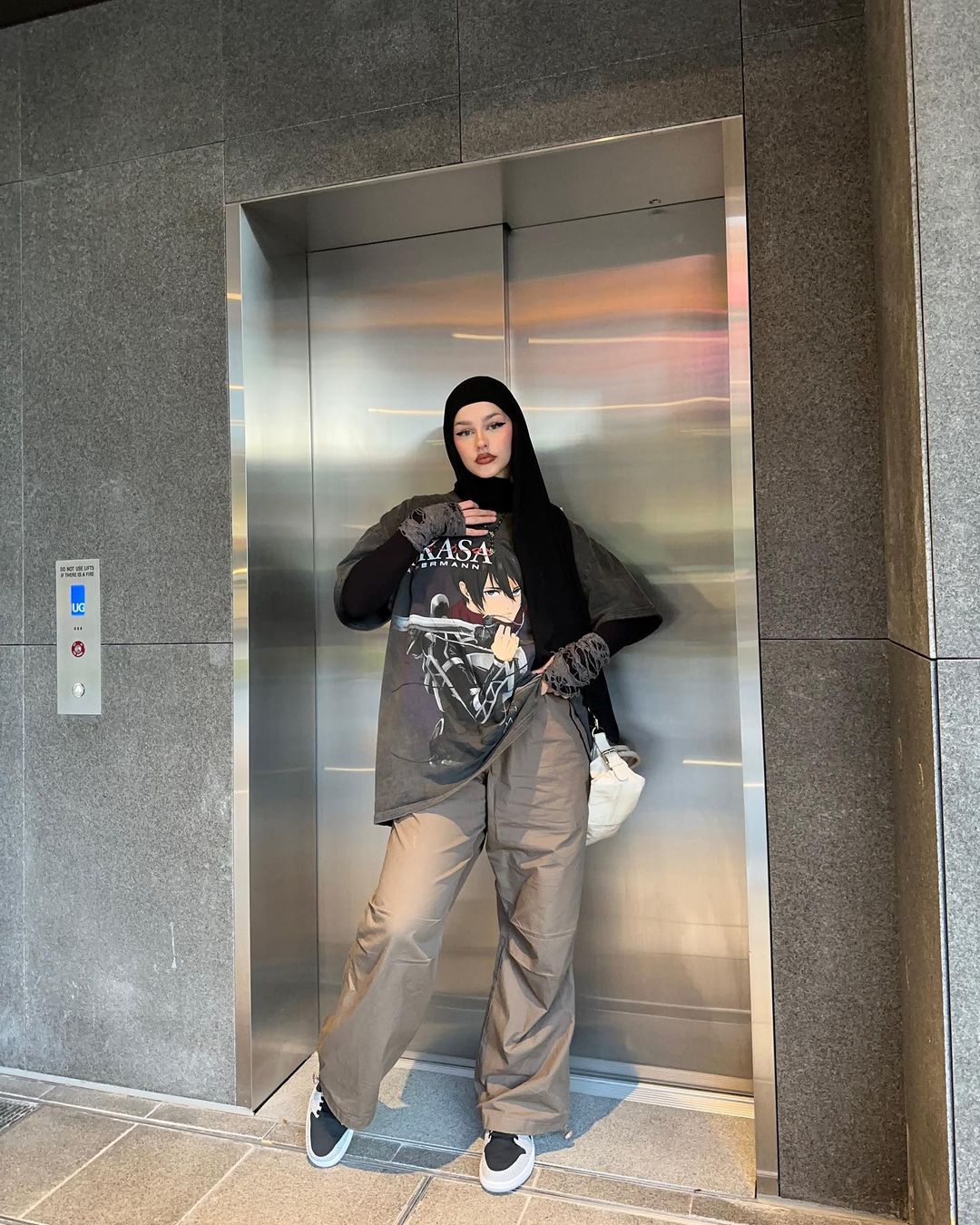 Stylish adult female posing in front of an elevator with a graphic tee, baggy pants, sneakers, and black headscarf indoors.