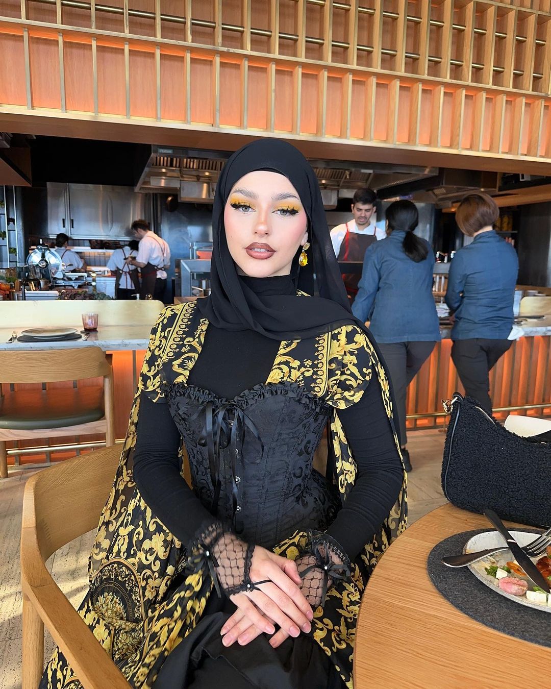 A woman wearing a black hijab and a black and gold patterned dress sits at a table in a restaurant with an open kitchen.