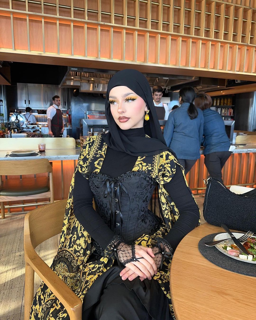 Woman in ornate black and gold corset dress and hijab sitting at a table in a restaurant. Indoor setting with other people in the background.