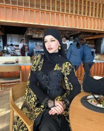 A woman wearing a black hijab and ornate gold and black patterned coat seated at a wooden table in a restaurant interior.