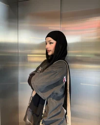 Woman in dark hijab and oversized grey t-shirt standing inside an elevator with metallic paneling