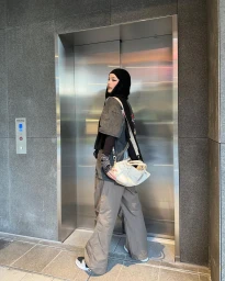 Woman in hijab wearing grey t-shirt, loose trousers, and carrying a white handbag standing by elevator.