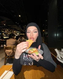 Woman in a black hijab holding a small food plate with tacos while seated at a wooden restaurant table