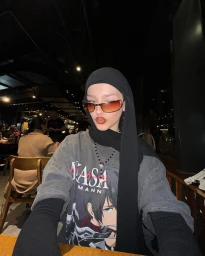 Woman wearing a black hijab and tinted sunglasses, styled with an oversized graphic t-shirt in a dimly lit venue.
