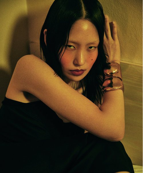 Asian woman with dark hair and bronze cuff poses indoors with dramatic makeup and flush while leaning against a neutral surface.