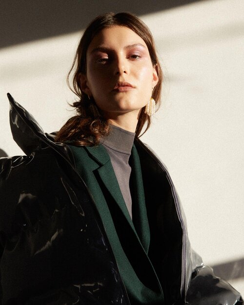 Fashion portrait of a woman in a dark green blazer and black patent leather jacket, posing in bright, diffused light against a neutral background.