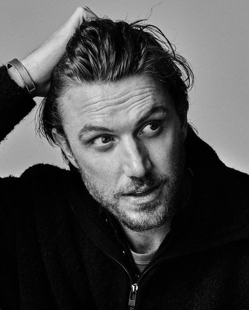 Black and white portrait of a man with his hand in his hair, wearing a coat, looking off to the side, showcasing a moody and thoughtful expression.