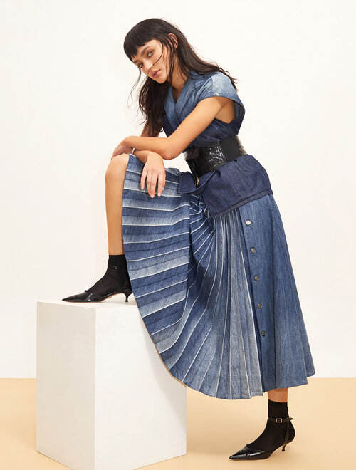 A woman poses in a denim kimono-style top and pleated skirt, leaning on a white block in a studio setting, showcasing a high-fashion look.