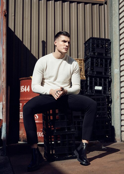 A young adult male poses while sitting on crates outdoors in a cream sweater, black pants, and black boots with an industrial background.