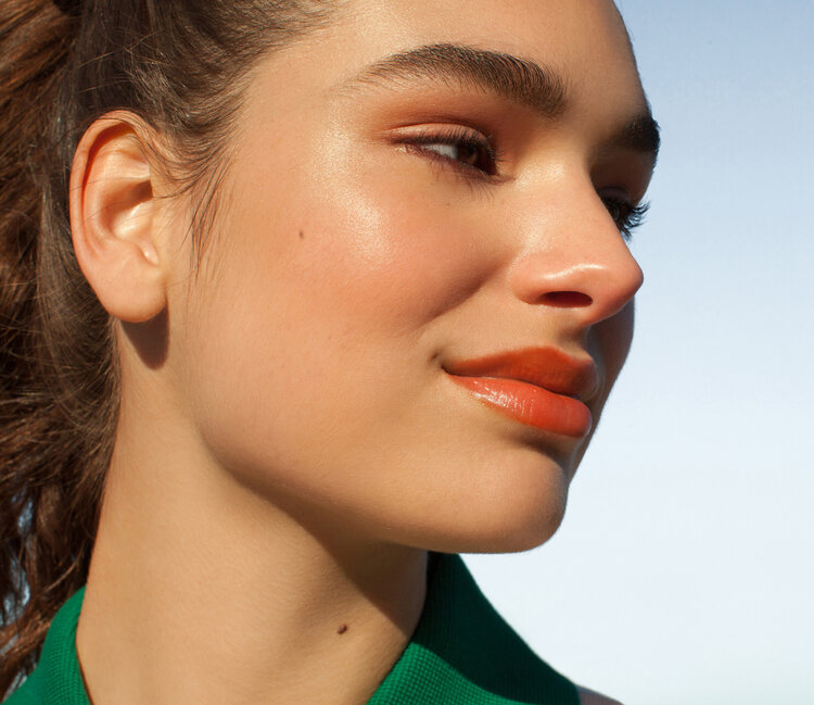 Close-up profile of a young adult female with brown hair in a ponytail, wearing a green top, against a bright blue sky background.
