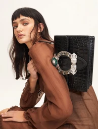 Model with blunt bangs wearing a sheer brown top holding a black crystal-buckled handbag against a white background