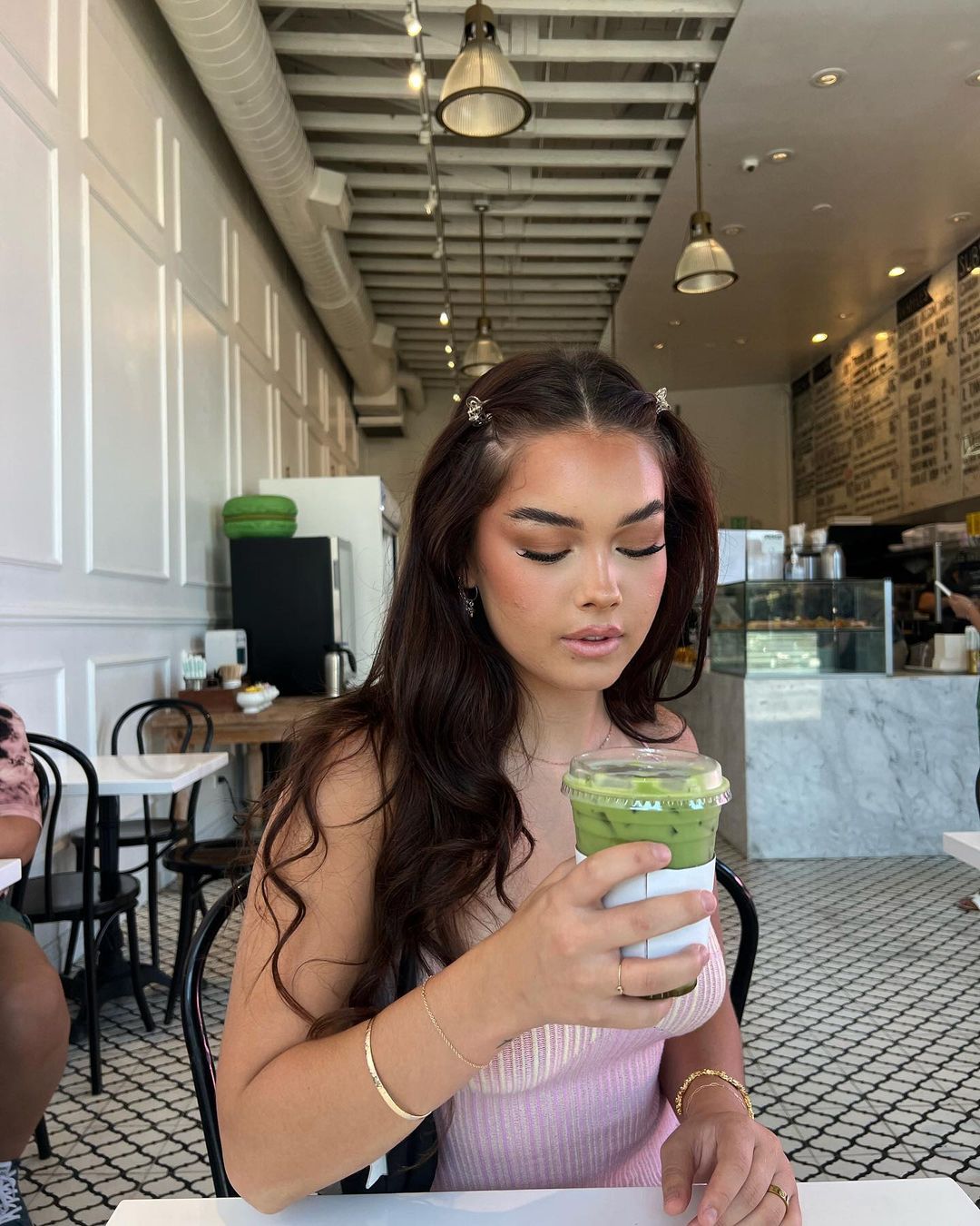 Young woman at a cafe table holds a green matcha latte. She wears a pink top, gold jewelry and decorative butterfly hair clips.