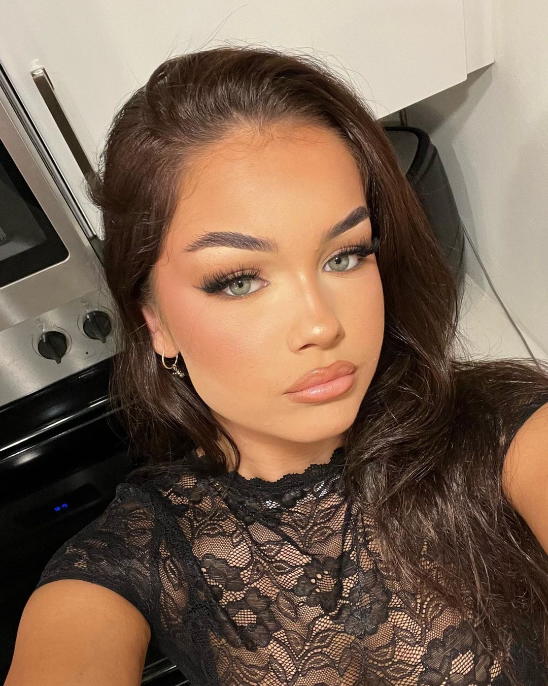 Close-up of a young woman with long brunette hair wearing a black lace top, posing in an indoor setting with muted lighting.