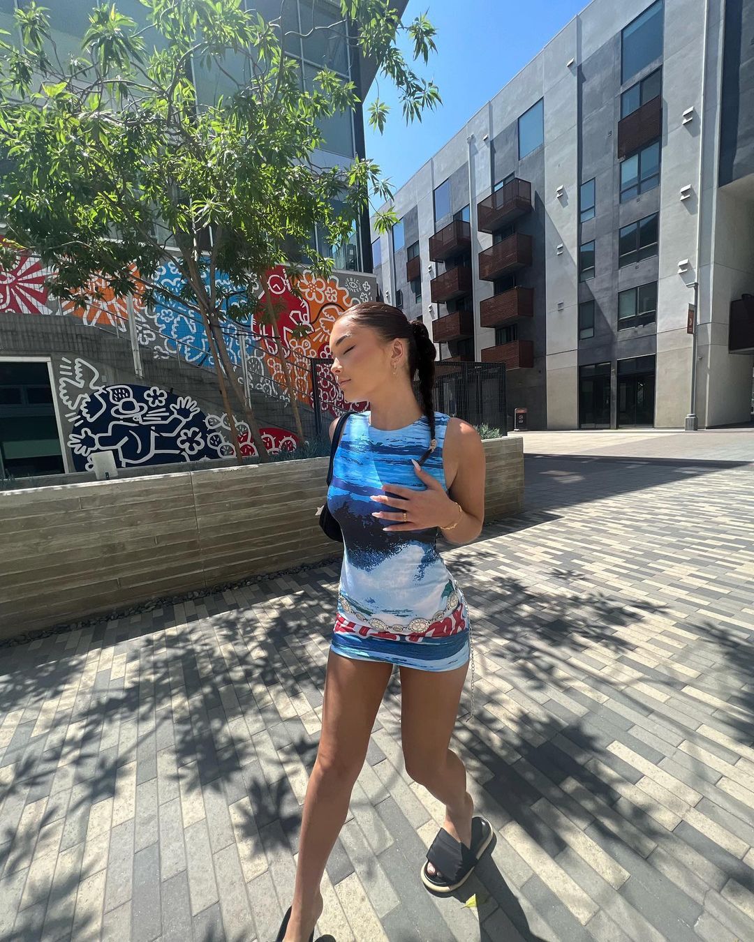 Woman in blue dress walks on brick pathway outdoors, shadowed by trees near a modern building with colorful mural art.