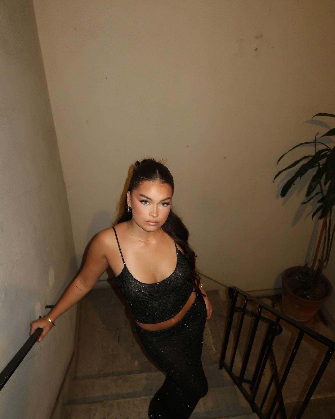 A young adult woman stands on a staircase wearing a glittering, two-piece outfit. She holds onto the railing and poses for the camera.