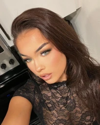 Close-up selfie of a woman with long dark hair wearing a black lace top in an indoor kitchen setting.