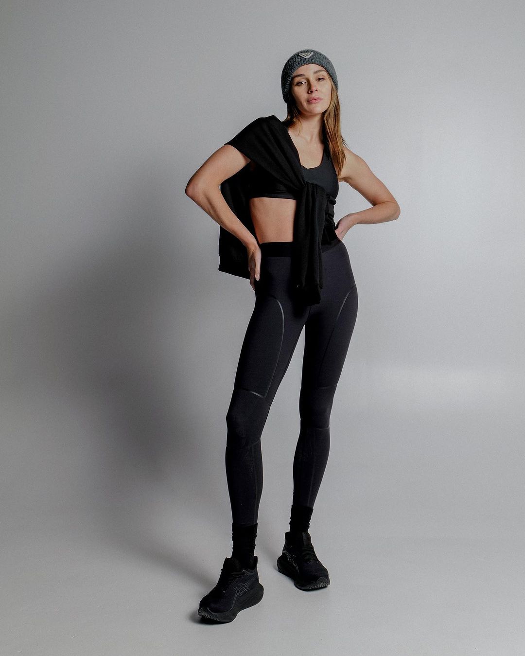 Full-length portrait of a fit adult woman in sportswear posing against a studio backdrop. She wears a beanie and athletic shoes.