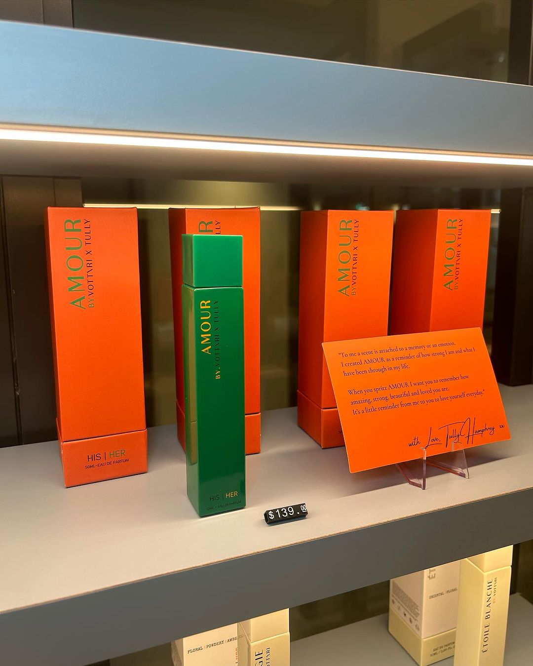 Shelf with green Amour perfume bottle and orange product boxes. Perfume bottle displayed in front of packaging.